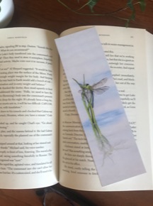 A realistic painting of a dragonfly reproduced on a bookmark sitting on a page of a book. 